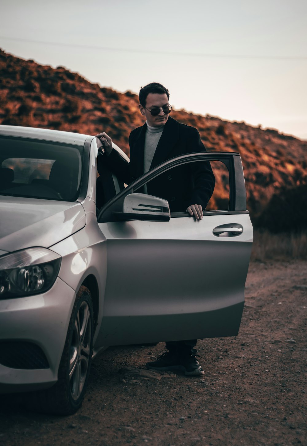 a man standing next to a silver car on a dirt road