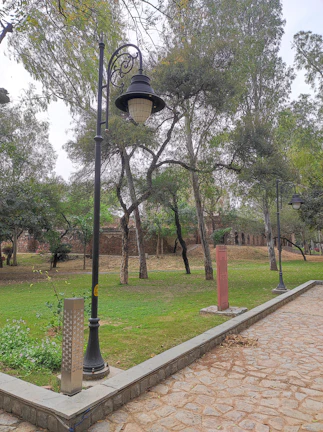 A park scene with a cobblestone pathway bordered by grass and trees. There are several tall black lampposts and informational signs positioned along the path. Green trees fill the background, creating a tranquil and natural environment.