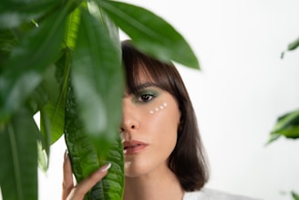 A woman's face partially obscured by large green leaves. She has brunette hair and green eyeshadow, with small white dots painted along her cheekbone and below her eye. She gazes directly at the camera in a serene manner.