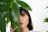 A woman's face partially obscured by large green leaves. She has brunette hair and green eyeshadow, with small white dots painted along her cheekbone and below her eye. She gazes directly at the camera in a serene manner.