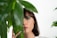 A woman's face partially obscured by large green leaves. She has brunette hair and green eyeshadow, with small white dots painted along her cheekbone and below her eye. She gazes directly at the camera in a serene manner.