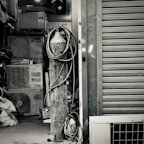 An industrial setting featuring a collection of stacked air conditioning units from various brands. A tall cylinder with hoses wrapped around it stands prominently in the foreground. The setting appears to be a cluttered storage or workshop space, with a partially visible metal roller shutter door on one side.