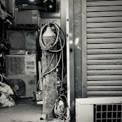 An industrial setting featuring a collection of stacked air conditioning units from various brands. A tall cylinder with hoses wrapped around it stands prominently in the foreground. The setting appears to be a cluttered storage or workshop space, with a partially visible metal roller shutter door on one side.