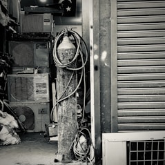 An industrial setting featuring a collection of stacked air conditioning units from various brands. A tall cylinder with hoses wrapped around it stands prominently in the foreground. The setting appears to be a cluttered storage or workshop space, with a partially visible metal roller shutter door on one side.