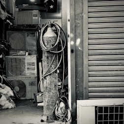An industrial setting featuring a collection of stacked air conditioning units from various brands. A tall cylinder with hoses wrapped around it stands prominently in the foreground. The setting appears to be a cluttered storage or workshop space, with a partially visible metal roller shutter door on one side.