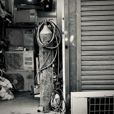An industrial setting featuring a collection of stacked air conditioning units from various brands. A tall cylinder with hoses wrapped around it stands prominently in the foreground. The setting appears to be a cluttered storage or workshop space, with a partially visible metal roller shutter door on one side.