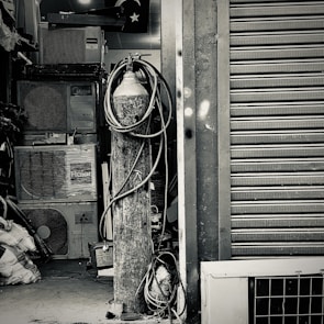 An industrial setting featuring a collection of stacked air conditioning units from various brands. A tall cylinder with hoses wrapped around it stands prominently in the foreground. The setting appears to be a cluttered storage or workshop space, with a partially visible metal roller shutter door on one side.