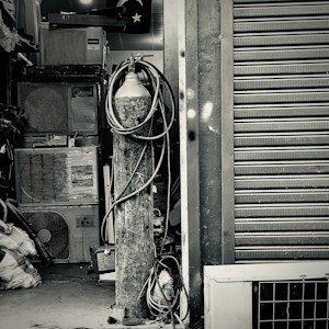 An industrial setting featuring a collection of stacked air conditioning units from various brands. A tall cylinder with hoses wrapped around it stands prominently in the foreground. The setting appears to be a cluttered storage or workshop space, with a partially visible metal roller shutter door on one side.