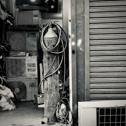 An industrial setting featuring a collection of stacked air conditioning units from various brands. A tall cylinder with hoses wrapped around it stands prominently in the foreground. The setting appears to be a cluttered storage or workshop space, with a partially visible metal roller shutter door on one side.