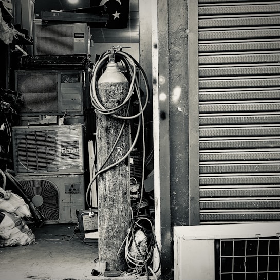 An industrial setting featuring a collection of stacked air conditioning units from various brands. A tall cylinder with hoses wrapped around it stands prominently in the foreground. The setting appears to be a cluttered storage or workshop space, with a partially visible metal roller shutter door on one side.