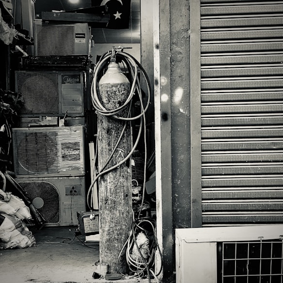 An industrial setting featuring a collection of stacked air conditioning units from various brands. A tall cylinder with hoses wrapped around it stands prominently in the foreground. The setting appears to be a cluttered storage or workshop space, with a partially visible metal roller shutter door on one side.