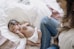 A newborn baby sleeps peacefully on a white pillow, wearing only a diaper. Next to the baby, a woman in blue jeans sits nearby, watching over the baby with gentle attention. The setting appears cozy and intimate, with soft fabrics around.