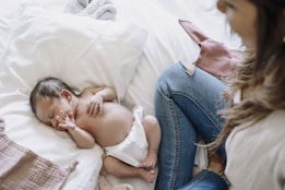 A newborn baby sleeps peacefully on a white pillow, wearing only a diaper. Next to the baby, a woman in blue jeans sits nearby, watching over the baby with gentle attention. The setting appears cozy and intimate, with soft fabrics around.
