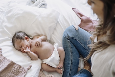 A newborn baby sleeps peacefully on a white pillow, wearing only a diaper. Next to the baby, a woman in blue jeans sits nearby, watching over the baby with gentle attention. The setting appears cozy and intimate, with soft fabrics around.