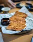 A plate of crispy, golden-brown fried food sits on a piece of branded paper. Accompanying the dish is a small, dark bowl filled with sauce. In the background, there's a blurred view of a hand reaching for something and other dishes on a wooden table.