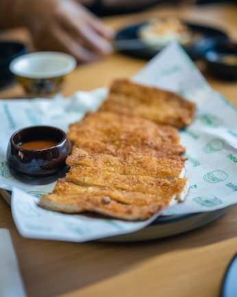 A plate of crispy, golden-brown fried food sits on a piece of branded paper. Accompanying the dish is a small, dark bowl filled with sauce. In the background, there's a blurred view of a hand reaching for something and other dishes on a wooden table.