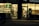 A warmly lit shop front is visible, with a person wearing a winter jacket standing in the entrance. The shop appears to be a mobile phone store, displaying phone accessories and offering services such as money transfer. An adjacent establishment, possibly a salon or barber shop, is visible with hair products displayed.