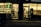 A warmly lit shop front is visible, with a person wearing a winter jacket standing in the entrance. The shop appears to be a mobile phone store, displaying phone accessories and offering services such as money transfer. An adjacent establishment, possibly a salon or barber shop, is visible with hair products displayed.