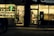 A warmly lit shop front is visible, with a person wearing a winter jacket standing in the entrance. The shop appears to be a mobile phone store, displaying phone accessories and offering services such as money transfer. An adjacent establishment, possibly a salon or barber shop, is visible with hair products displayed.