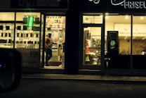 A warmly lit shop front is visible, with a person wearing a winter jacket standing in the entrance. The shop appears to be a mobile phone store, displaying phone accessories and offering services such as money transfer. An adjacent establishment, possibly a salon or barber shop, is visible with hair products displayed.