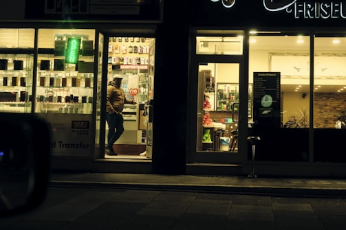 A warmly lit shop front is visible, with a person wearing a winter jacket standing in the entrance. The shop appears to be a mobile phone store, displaying phone accessories and offering services such as money transfer. An adjacent establishment, possibly a salon or barber shop, is visible with hair products displayed.