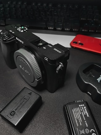 A digital camera, likely a Sony model, is placed on a dark surface. Accompanying items include camera batteries, a battery charger, and a red smartphone. A keyboard is visible in the background, suggesting a workspace environment.