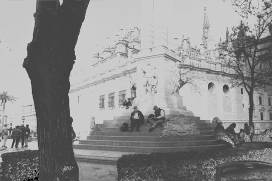 A black and white photograph depicts a historic building with ornate architectural details in the background. In the foreground, a group of people sit on the steps of a monument, which features statues. The scene is framed by trees on either side, creating a contrast between the natural elements and the constructed environment.