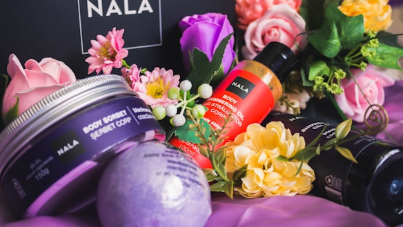 An arrangement of beauty products surrounded by colorful flowers. The focus is on several Nala brand items, including body sorbet and body oil, alongside various floral elements such as pink, purple, and yellow blossoms. The background is a dark surface that enhances the vibrancy of the flowers and products.
