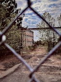 An abandoned building is partially obscured by a chain-link fence with barbed wire on top. The structure has multiple levels with red and white accents and is surrounded by overgrown vegetation and trees. The sky is partly cloudy, providing a backdrop that adds to the deserted and neglected atmosphere.