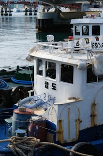 A small, weathered fishing boat is docked at a marina. The vessel shows signs of rust and wear, and there are various fishing paraphernalia like ropes, barrels, and hoses on deck. In the background, a concrete bridge and other boats are visible on a calm body of water.