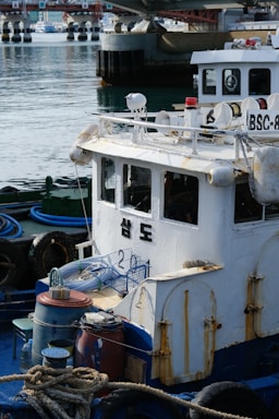 A small, weathered fishing boat is docked at a marina. The vessel shows signs of rust and wear, and there are various fishing paraphernalia like ropes, barrels, and hoses on deck. In the background, a concrete bridge and other boats are visible on a calm body of water.