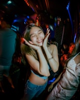 A young woman is smiling with her eyes closed in a lively party atmosphere. She is holding her hands to her face in a playful gesture. There is colorful lighting with neon effects, and people around are also enjoying the mood.