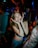 A young woman is smiling with her eyes closed in a lively party atmosphere. She is holding her hands to her face in a playful gesture. There is colorful lighting with neon effects, and people around are also enjoying the mood.
