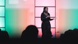 A person is standing on stage, engaging with an audience. The background is lit with colorful lighting, transitioning from green on the left to red on the right. The person appears to be speaking or presenting, with an expressive posture. The scene is viewed from the perspective of the audience, some of whom are silhouetted in the foreground.