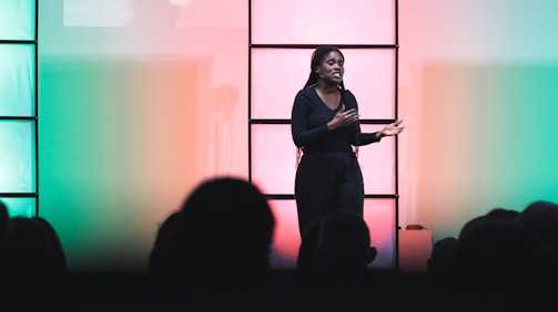 A person is standing on stage, engaging with an audience. The background is lit with colorful lighting, transitioning from green on the left to red on the right. The person appears to be speaking or presenting, with an expressive posture. The scene is viewed from the perspective of the audience, some of whom are silhouetted in the foreground.
