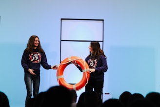 Two people are on a stage wearing matching dark hoodies with a logo. They are holding a large orange lifebuoy, smiling at each other. A simple backdrop with a light blue hue is in the background, and a partitioned panel stands behind them.