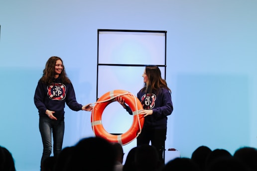 Two people are on a stage wearing matching dark hoodies with a logo. They are holding a large orange lifebuoy, smiling at each other. A simple backdrop with a light blue hue is in the background, and a partitioned panel stands behind them.