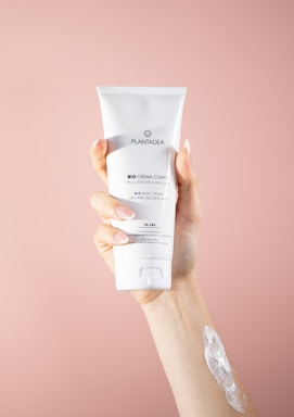 A hand holding a tube of white body cream labeled 'PLANTADEA Bio Crema Corpo' against a soft pink background. There is a dab of cream applied on the forearm of the hand holding the tube.
