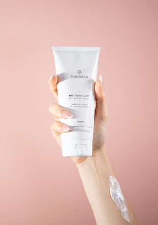 A hand holding a tube of white body cream labeled 'PLANTADEA Bio Crema Corpo' against a soft pink background. There is a dab of cream applied on the forearm of the hand holding the tube.