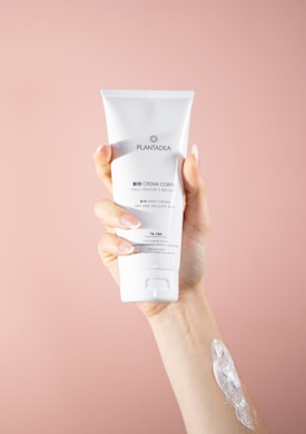 A hand holding a tube of white body cream labeled 'PLANTADEA Bio Crema Corpo' against a soft pink background. There is a dab of cream applied on the forearm of the hand holding the tube.