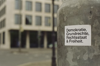 A sticker with German text 'Demokratie, Grundrechte, Rechtsstaat & Freiheit' is attached to a weathered metallic surface, likely a pole. The background features a blurred view of a city street and a modern building with multiple windows.