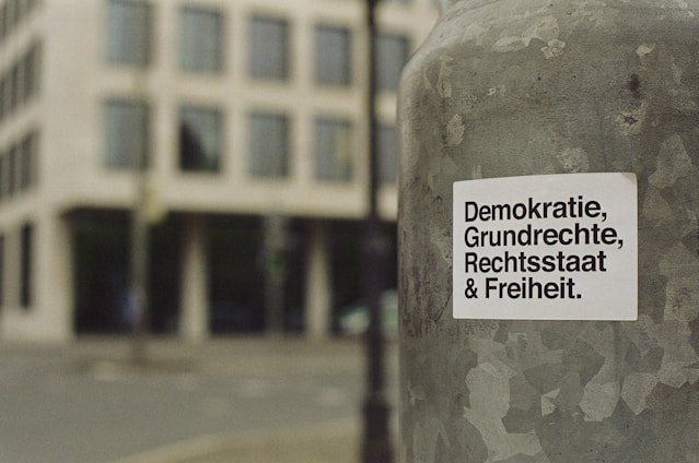 A sticker with German text 'Demokratie, Grundrechte, Rechtsstaat & Freiheit' is attached to a weathered metallic surface, likely a pole. The background features a blurred view of a city street and a modern building with multiple windows.