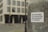 A sticker with German text 'Demokratie, Grundrechte, Rechtsstaat & Freiheit' is attached to a weathered metallic surface, likely a pole. The background features a blurred view of a city street and a modern building with multiple windows.
