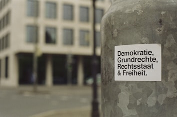 A sticker with German text 'Demokratie, Grundrechte, Rechtsstaat & Freiheit' is attached to a weathered metallic surface, likely a pole. The background features a blurred view of a city street and a modern building with multiple windows.