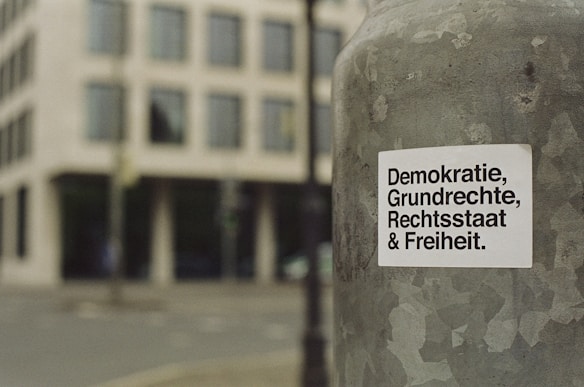 A sticker with German text 'Demokratie, Grundrechte, Rechtsstaat & Freiheit' is attached to a weathered metallic surface, likely a pole. The background features a blurred view of a city street and a modern building with multiple windows.