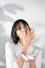 A person with short dark hair is applying cream to their face using their fingers. They are holding an open jar of skincare product in their other hand. The person is wearing a white shirt, and there is a soft focus shadow in the background.