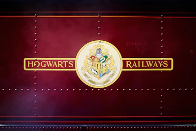 A rich burgundy background with a central gold and red emblem displaying the Hogwarts Railways crest, featuring symbols representing the four houses of the fictional Hogwarts School. The words 'Hogwarts Railways' are printed in an ornate style on either side of the emblem.