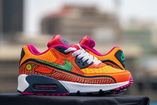 A pair of vibrant, multicolored sneakers with intricate patterns and mesh textures. The shoes feature a combination of orange, pink, green, and black with a white midsole and black outsole. There is a prominent swoosh and 'Air Max' branding visible.