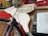 A bicycle features a unique wooden decoration of a flying pig attached to the front. The pig is painted in a light color with wings, suggesting a whimsical or recycled theme. Behind the bicycle, parts of a red and yellow structure are visible, adding a colorful background.