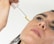 A close-up of a person applying a skincare serum or oil to their cheek using a dropper. The person has neutral expression with a focus on skincare and wellness.